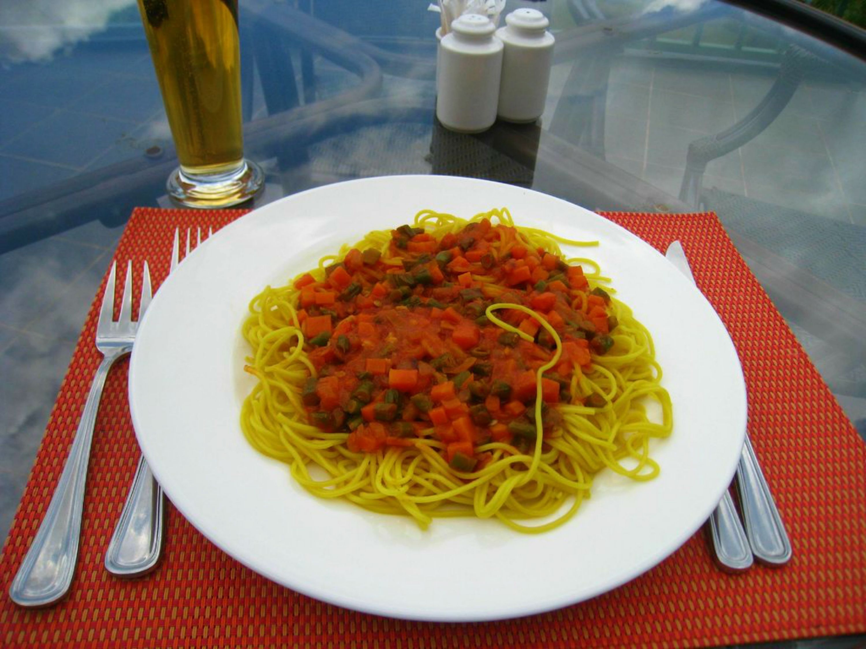 A plate of spaghetti with a tomato-based sauce, accompanied by a glass of beer and salt and pepper shakers on a glass table.