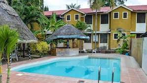 A swimming pool with a thatched umbrella, surrounded by palm trees and a yellow building with a red roof. - Ogalis K Coast Hotel (Mtwapa)