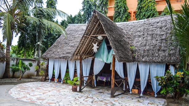 A thatched-roof hut with white curtains and a tiled floor, surrounded by palm trees and plants. - Ogalis K Coast Hotel (Mtwapa)