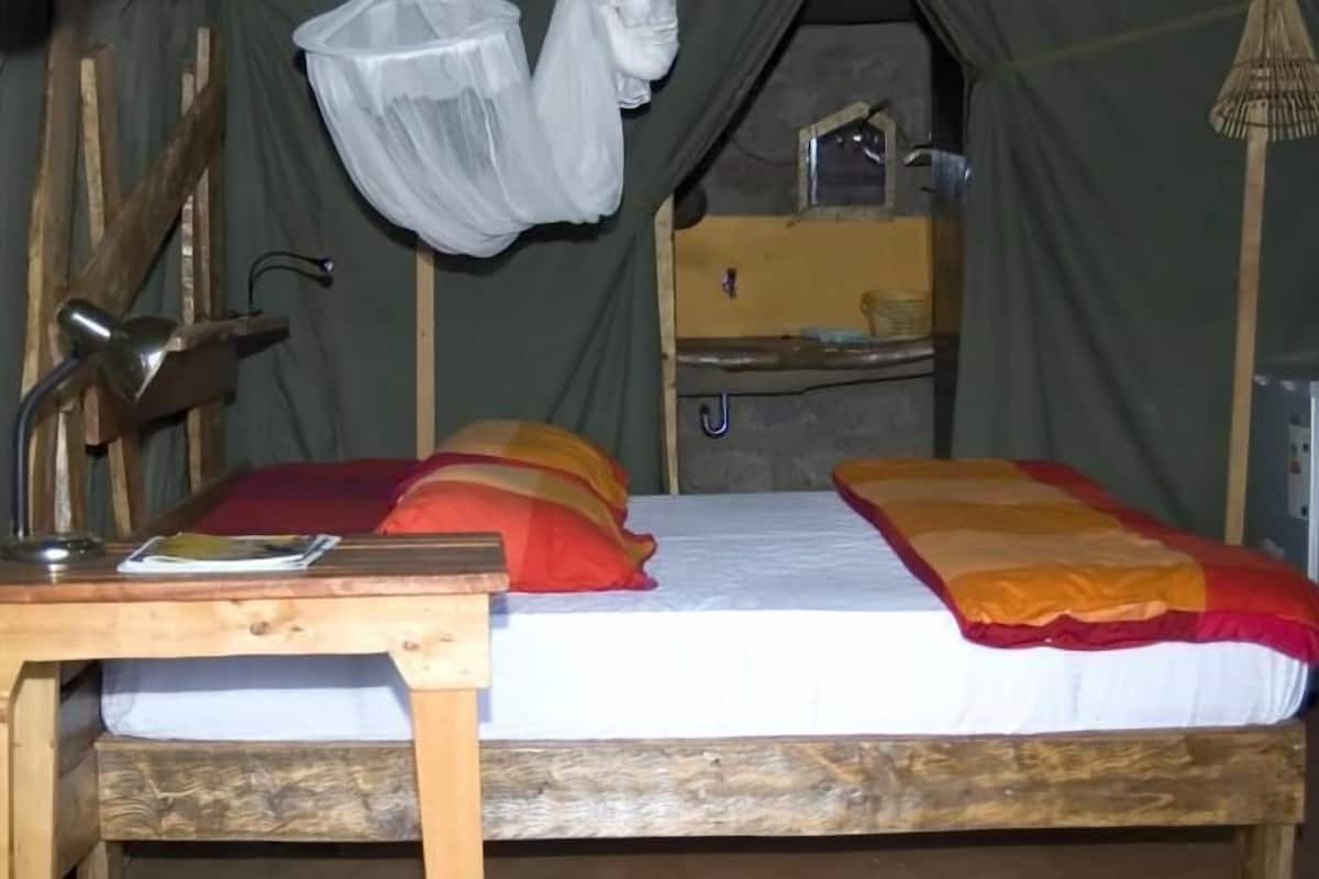 A wooden bed with a white mattress and colorful pillows, a small wooden table with a lamp, and a hanging mosquito net.
