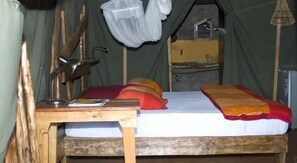 A wooden bed with a white mattress and colorful pillows, a small wooden table with a lamp, and a hanging mosquito net.