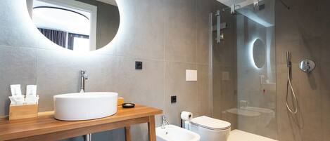 Een moderne badkamer met een ronde spiegel, een houten wastafelmeubel, een toilet, een douche met glazen wanden en een bidet.