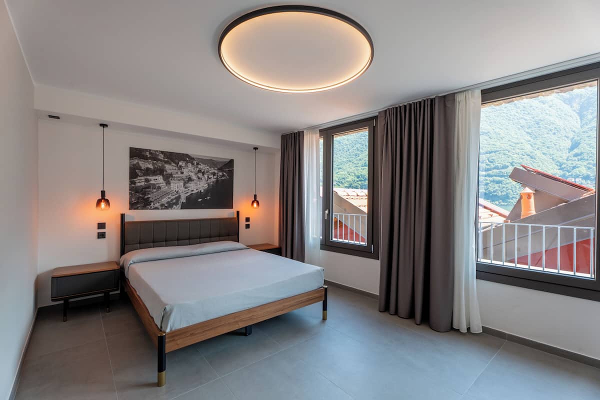 A modern bedroom with a bed, two bedside lamps, a ceiling light, a large window with curtains, and a cityscape painting on the wall.