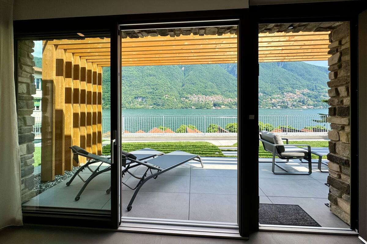 A modern room with a sliding glass door leading to a balcony with a view of a lake and mountains.