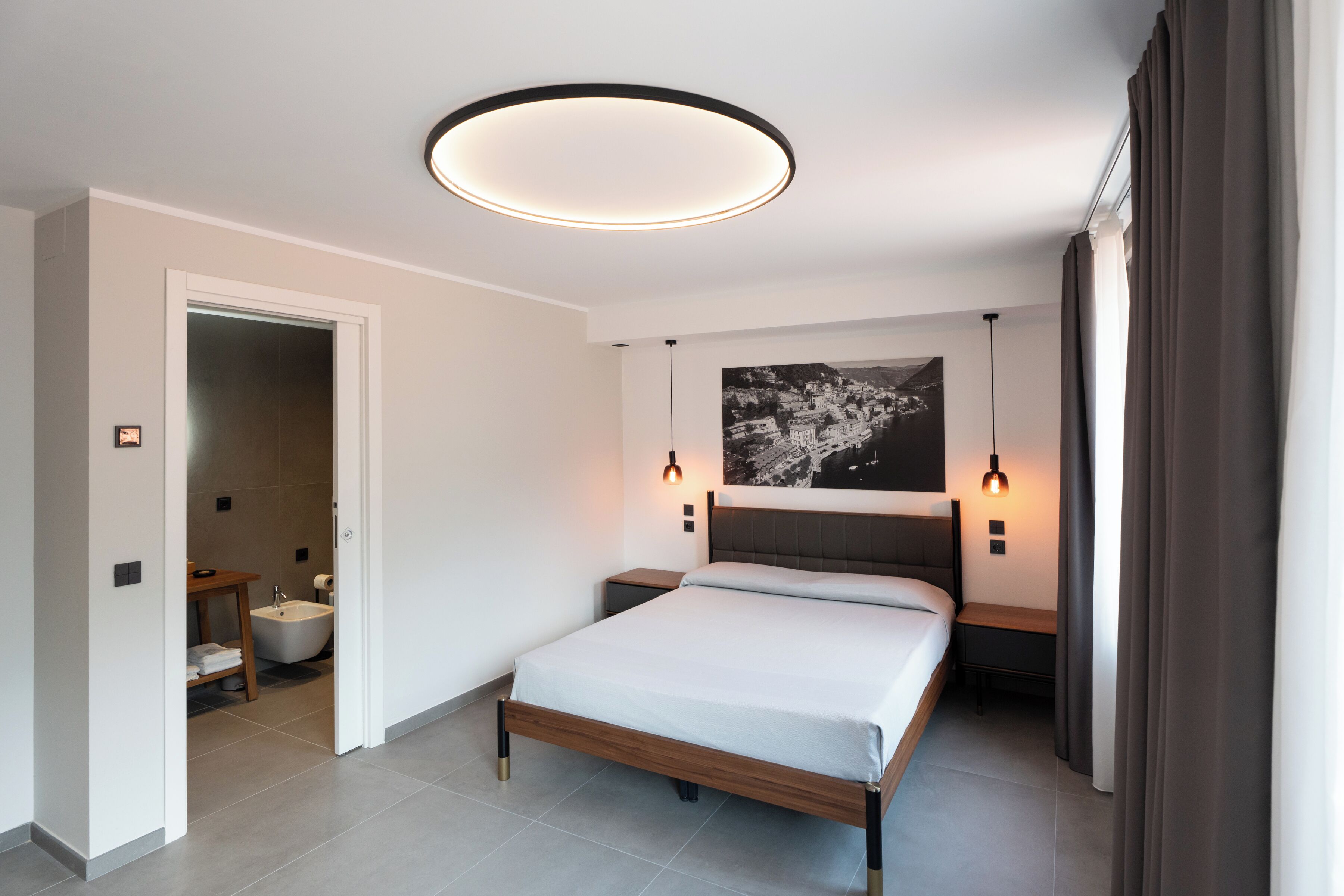Une chambre moderne avec un lit, des tables de chevet, une tête de lit, un luminaire fixé au mur et une salle de bain visible à travers une porte ouverte.