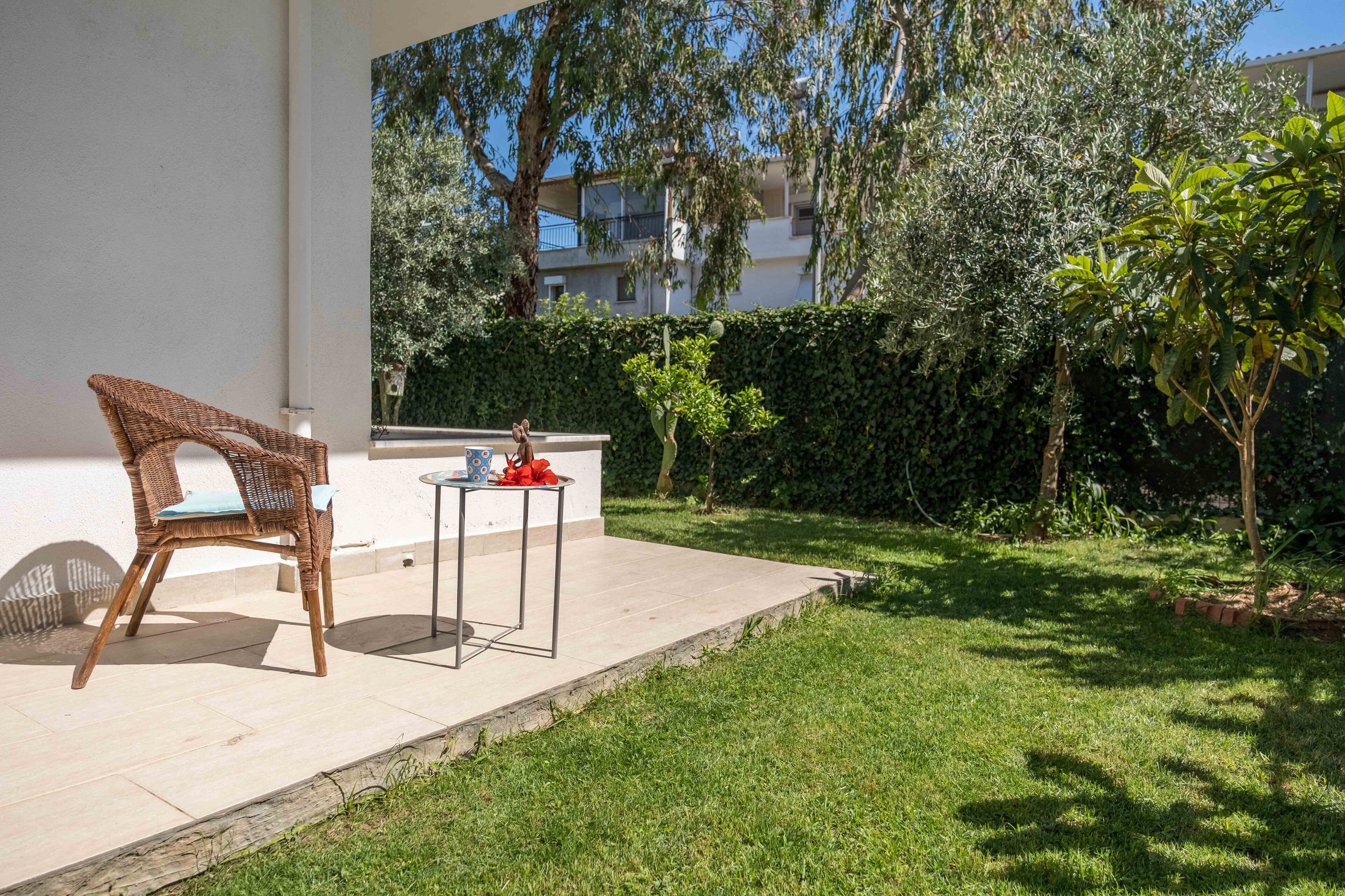 Un patio con una silla de mimbre y una mesita, rodeado de césped cuidado y setos.