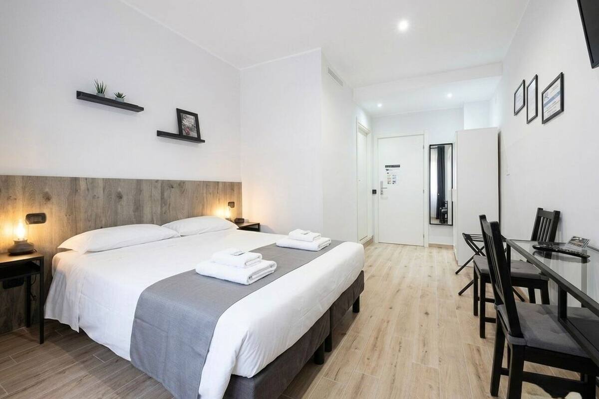 A modern hotel room with a large bed, bedside tables, a desk with chairs, and a mirror on the wall.