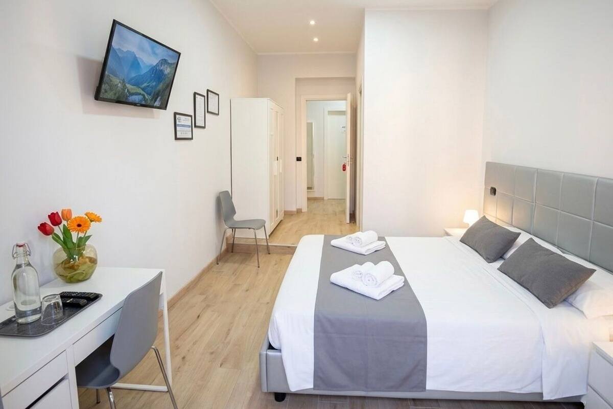 A hotel room with a bed, a desk with a chair, a TV mounted on the wall, and a vase with flowers on the desk.