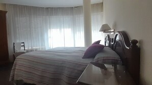 Quarto de hotel com cama, mesa de cabeceira, luminária e janela com cortinas translúcidas.
