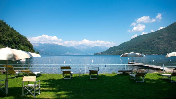 Vista de un lago con sillones de descanso y sombrillas, un muelle y montañas al fondo.