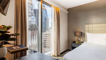 A hotel room with a bed, a bedside table with a lamp, a small table with a book, a potted plant, and a view of a cityscape through large windows.