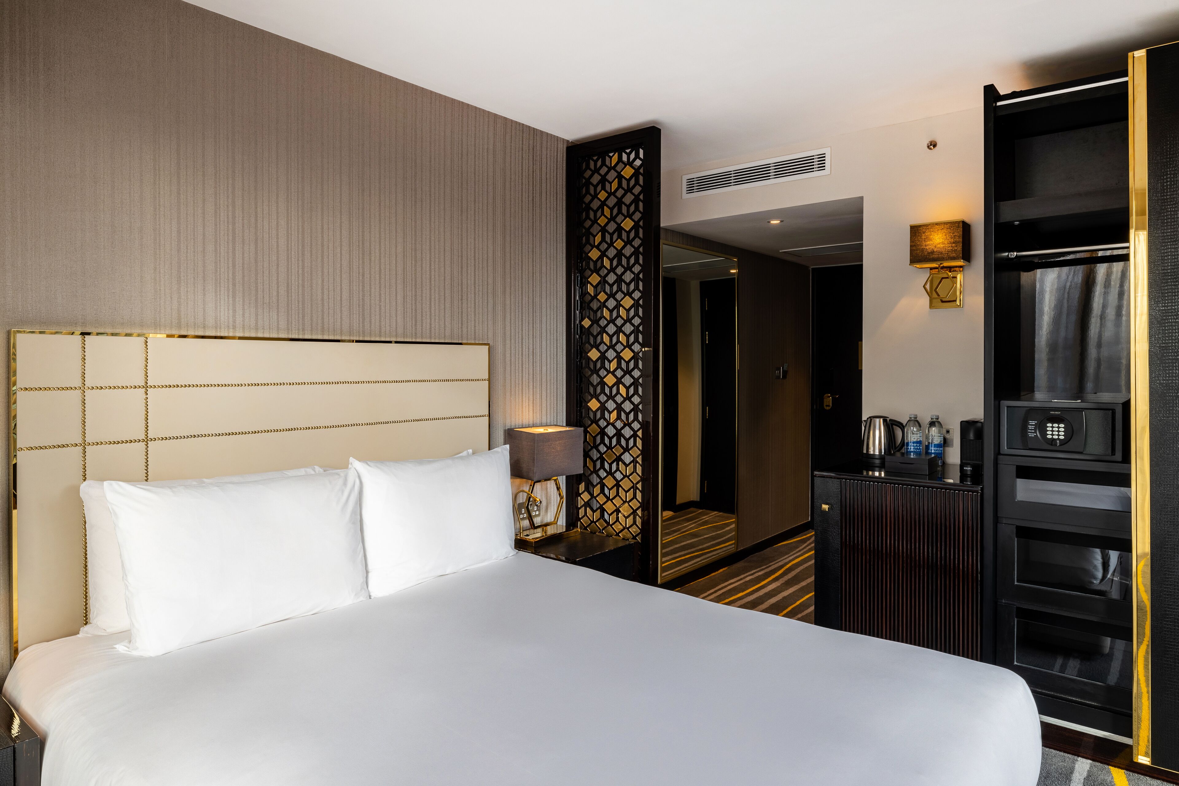A hotel room with a large bed, a desk with a lamp, a minibar, and a wardrobe.