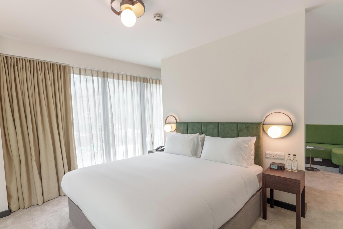Hotellihuone, jossa on suuri sänky, kaksi yöpöytävalaisinta, yöpöytä, jossa on puhelin ja pullo, sekä vihreä, verhoiltu pääty.