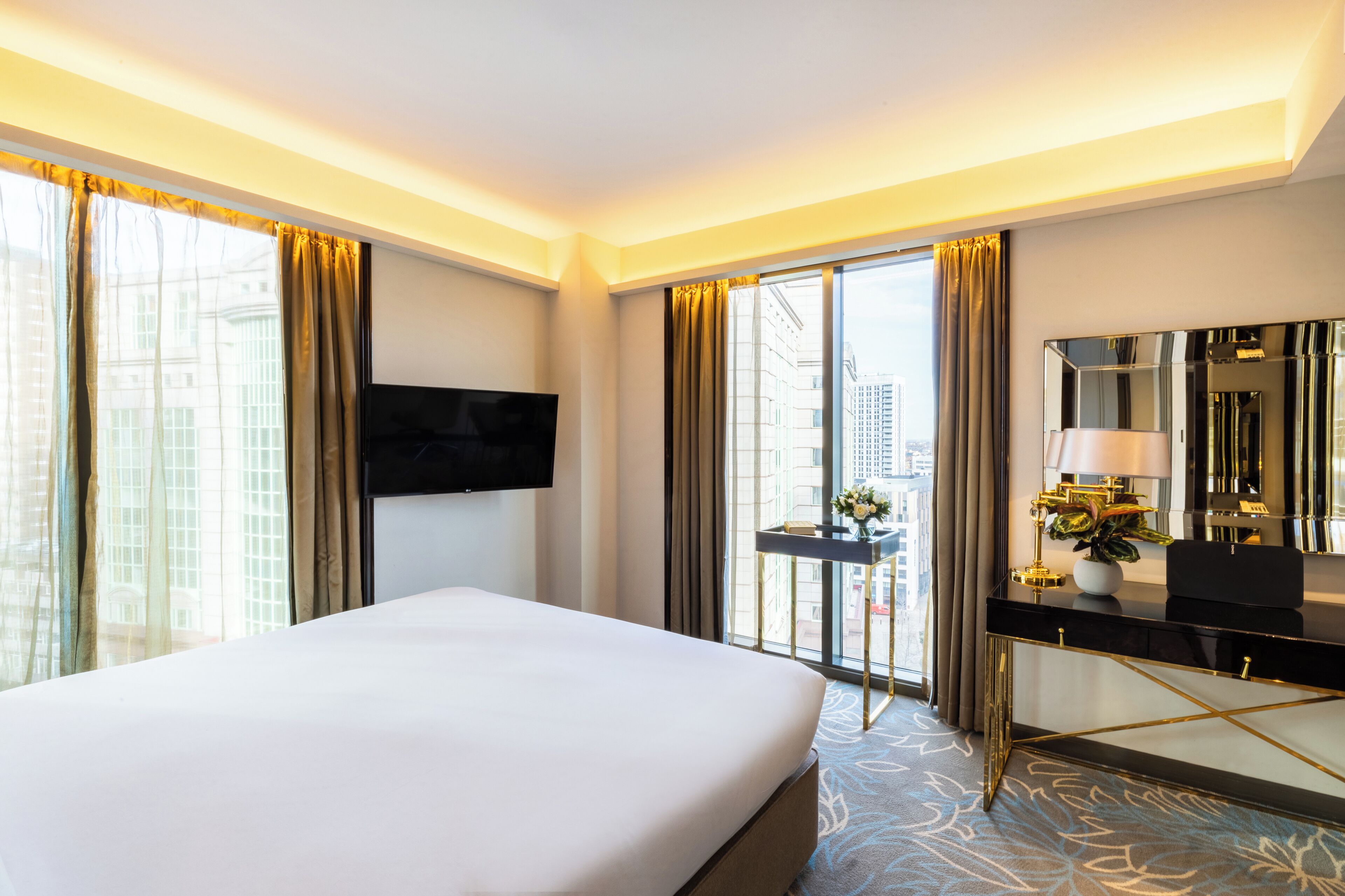 Ein Hotelzimmer mit Bett, Wandfernseher, Schreibtisch mit Lampe und großem Fenster mit Vorhängen.