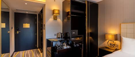 A hotel room with a bed, bedside table, lamp, and a desk with a microwave, coffee maker, and glasses.