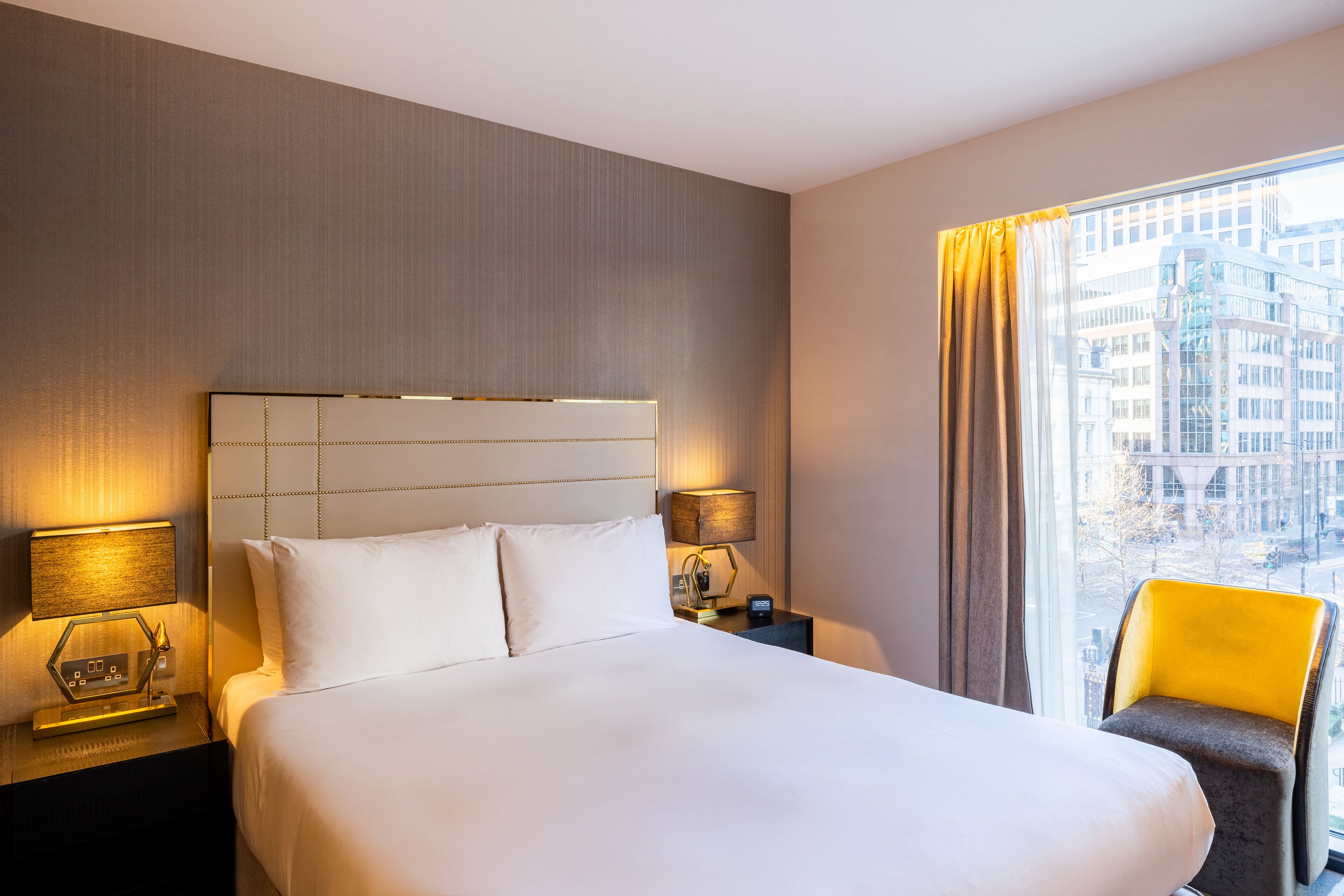 A hotel room with a large bed, two bedside lamps, a nightstand, a chair, and a window with a city view.
