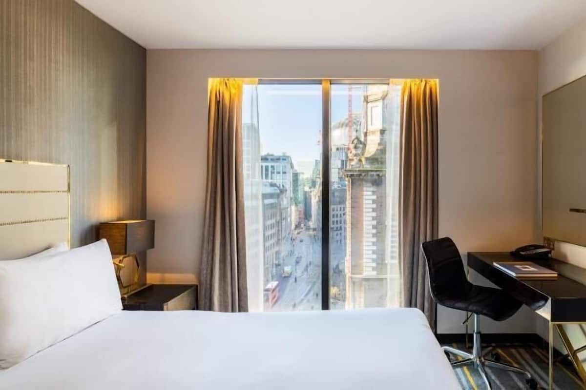 Una habitación de hotel con cama, escritorio, silla, televisor y un ventanal con cortinas.