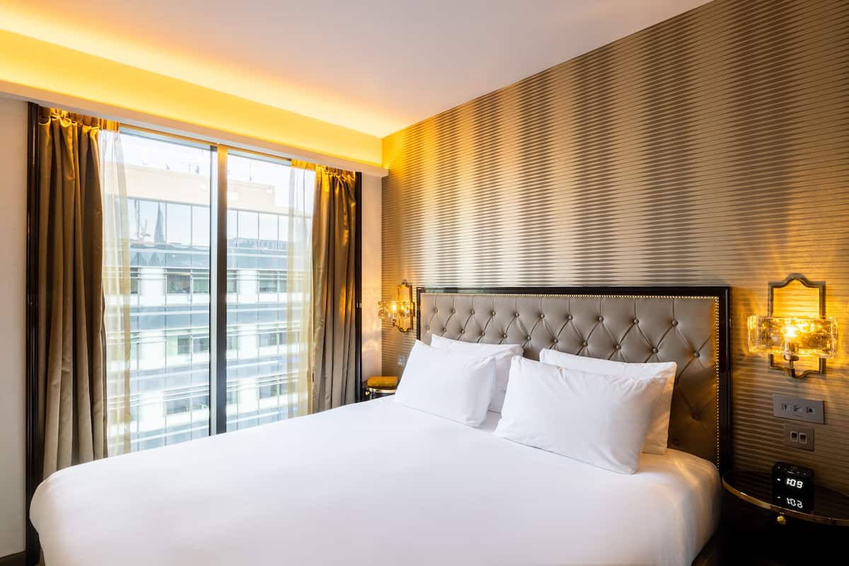 Et hotelværelse med en stor seng, en sengegavl med en quiltet detalje og udsigt over byen gennem gulv til loft-vinduer.