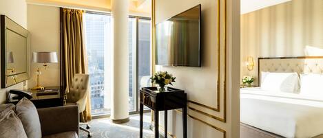 Quarto de hotel com uma cama grande, uma escrivaninha com abajur, uma cadeira, uma televisão e vista para a cidade.