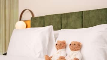 A bed with white pillows and a headboard, two teddy bears sitting on the pillows, and a bedside lamp.