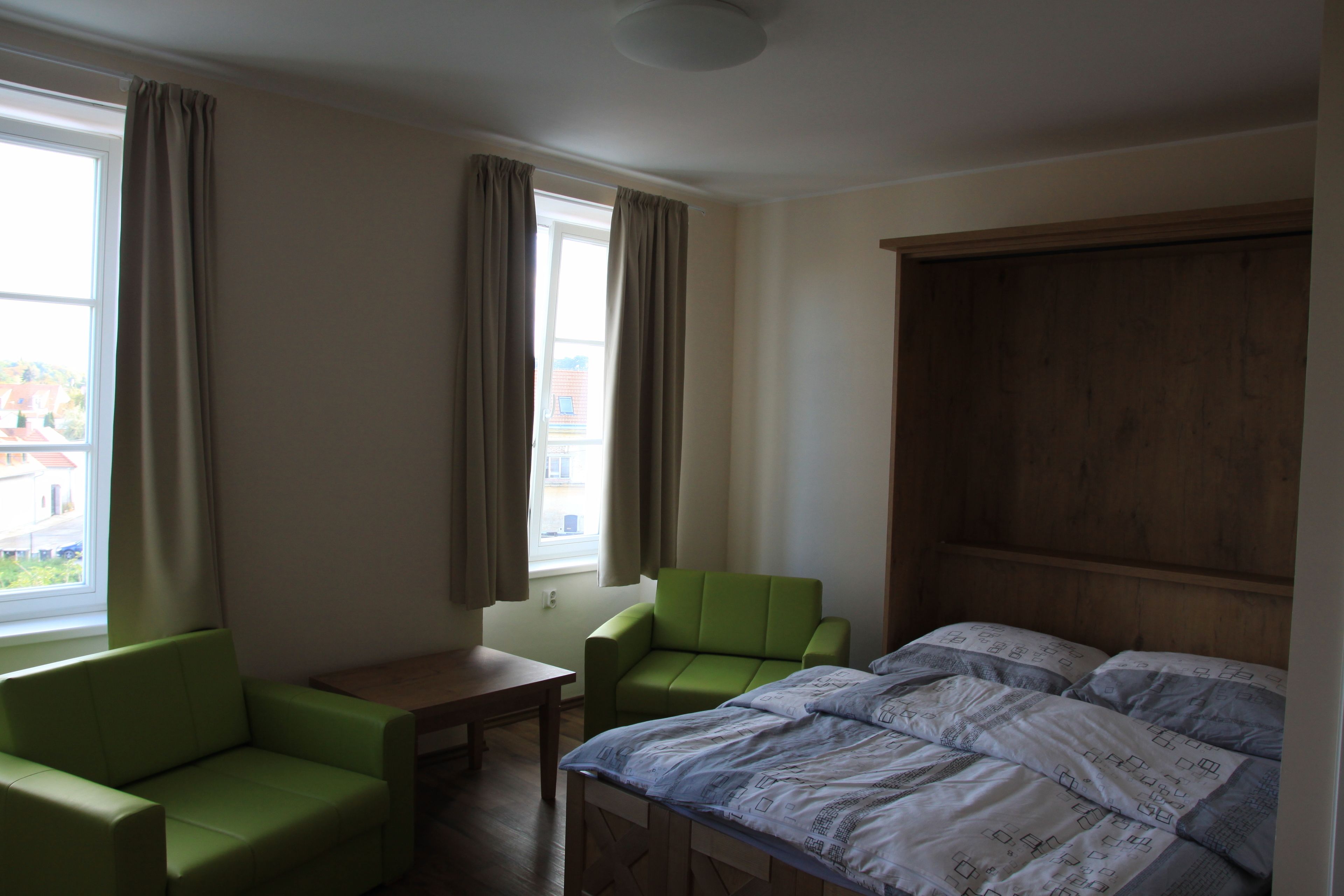 A hotel room with a bed, a green armchair, a wooden headboard, a small table, and a window with curtains.