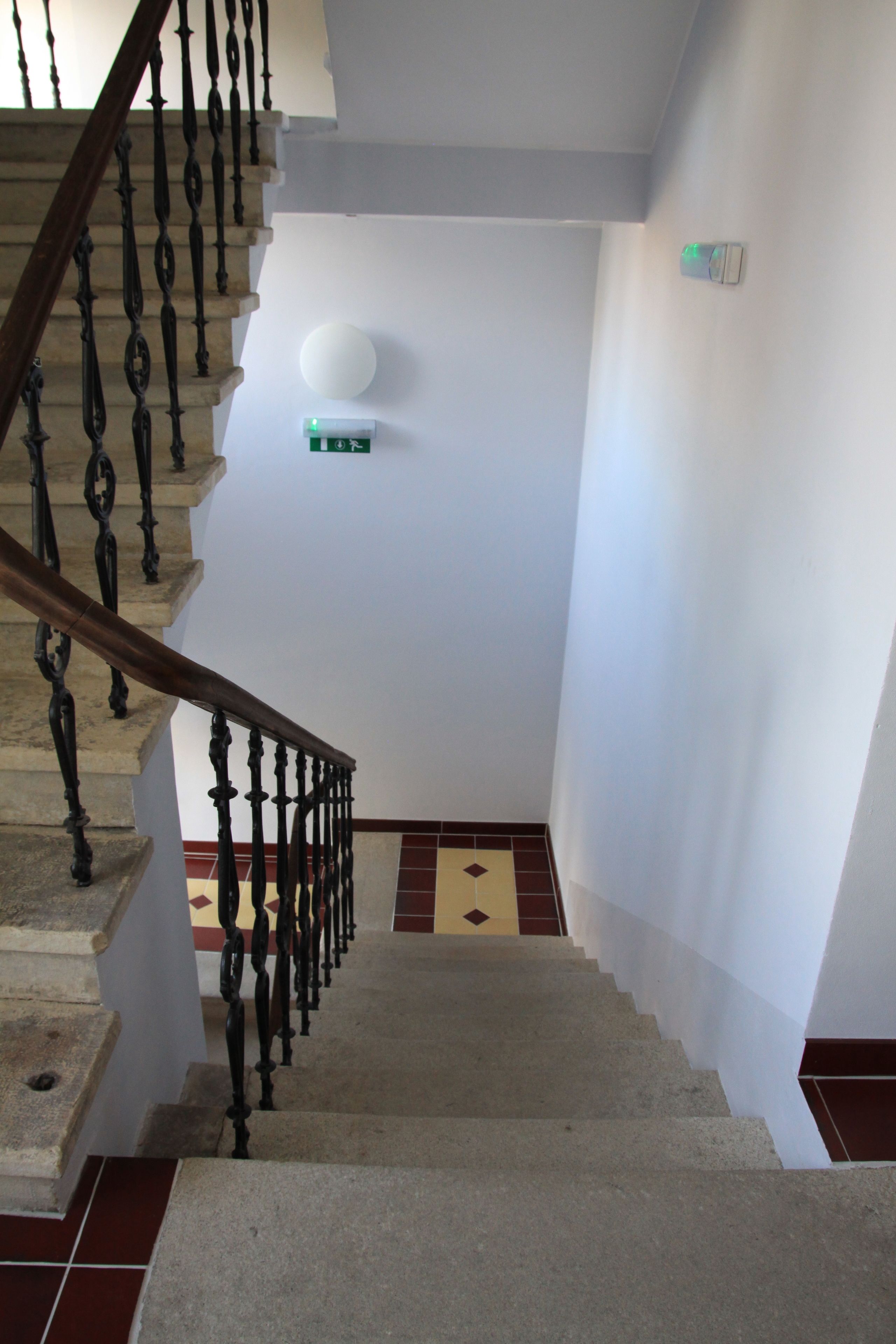 A staircase with a wooden handrail and black balusters, leading to a landing with a white wall and a green emergency exit sign.