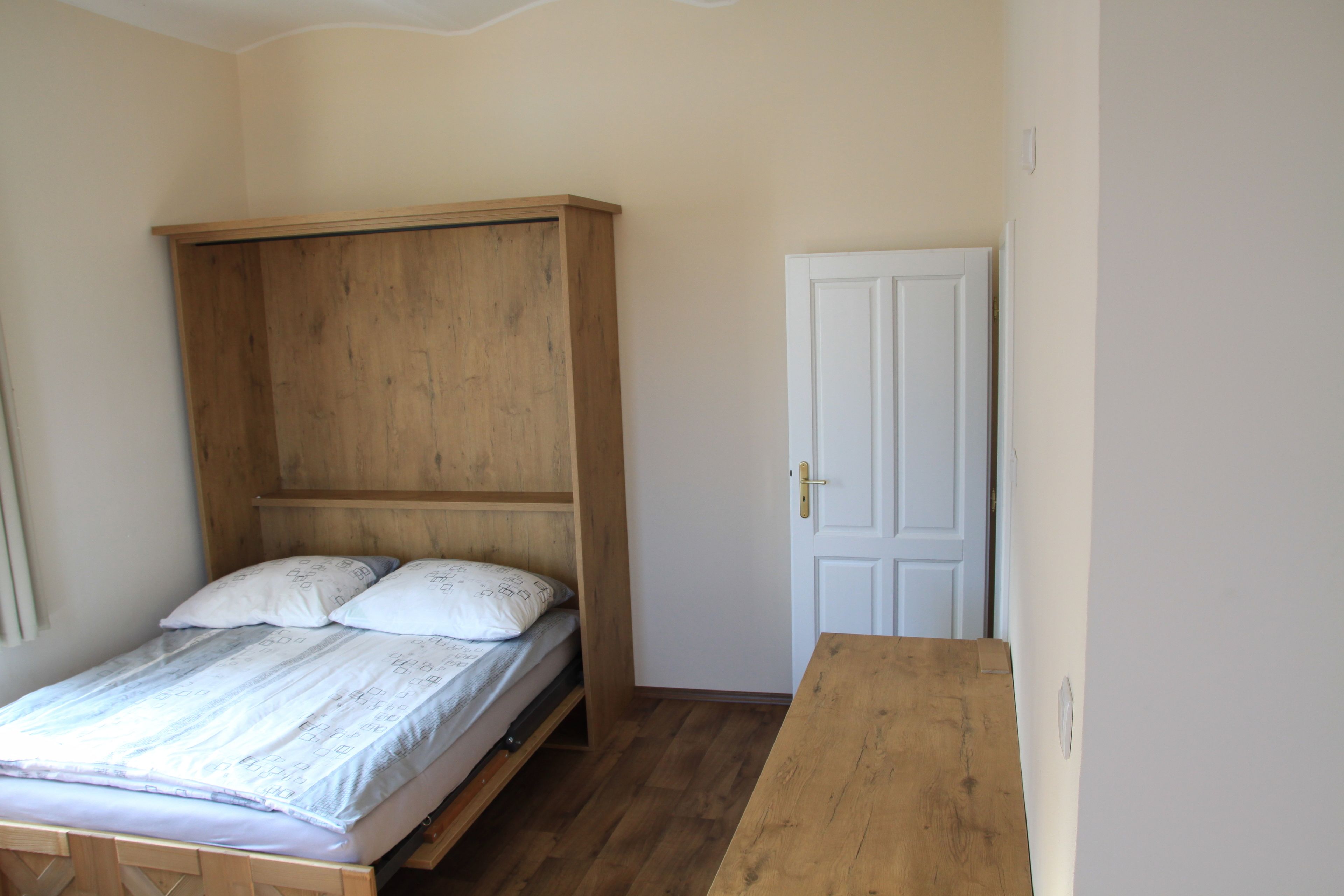 Ein kleines Zimmer mit einem Einzelbett, einem Holzkopfteil, einer weißen Tür und einem Holztisch.