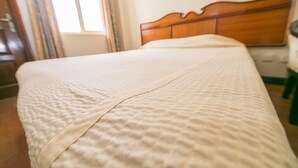 Uma cama com colcha branca texturizada num quarto com cabeceira de madeira e janela com cortinas.