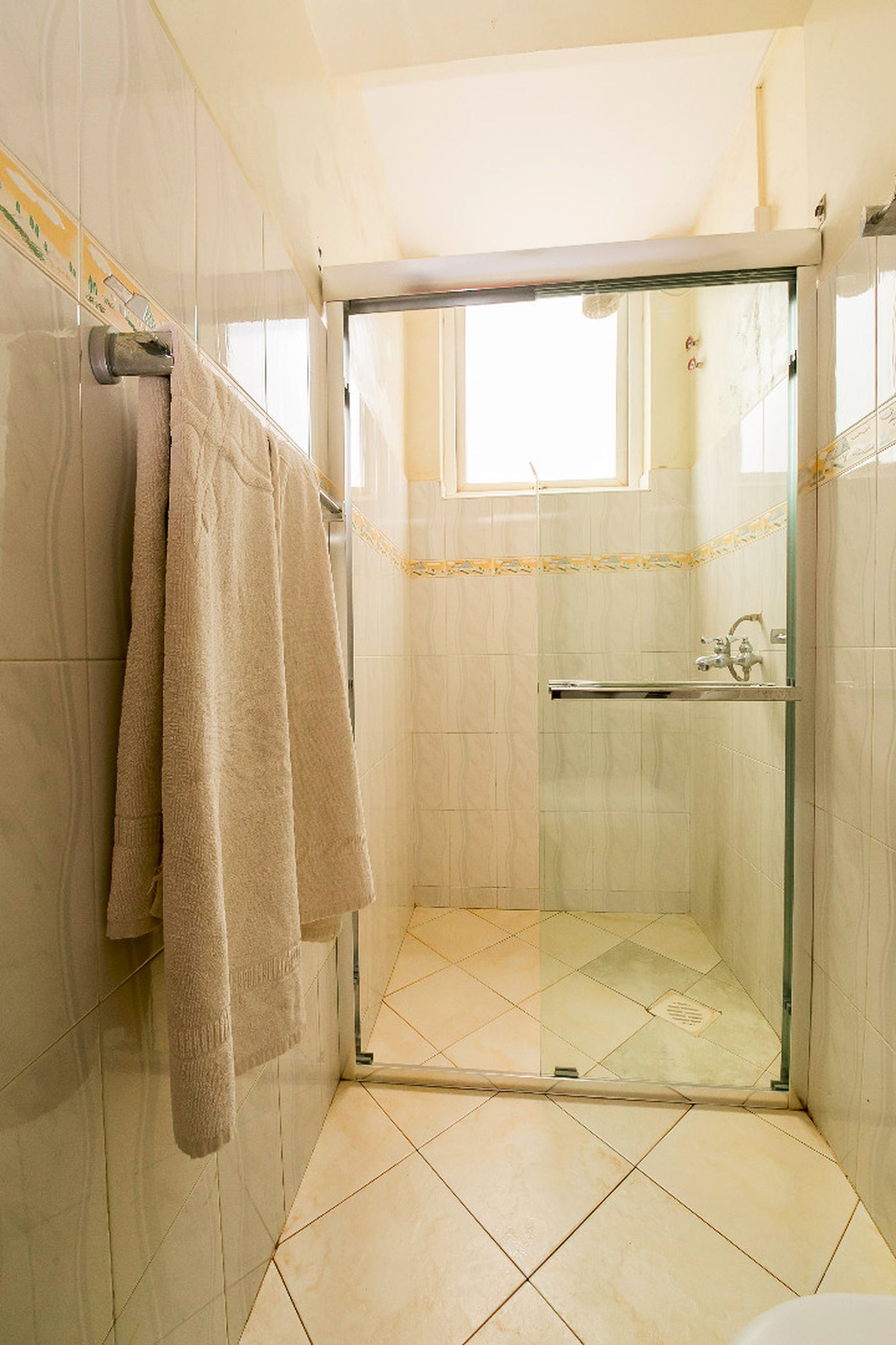 Ducha con paredes de vidrio y una pequeña ventana, un perchero con toallas y piso de baldosas.