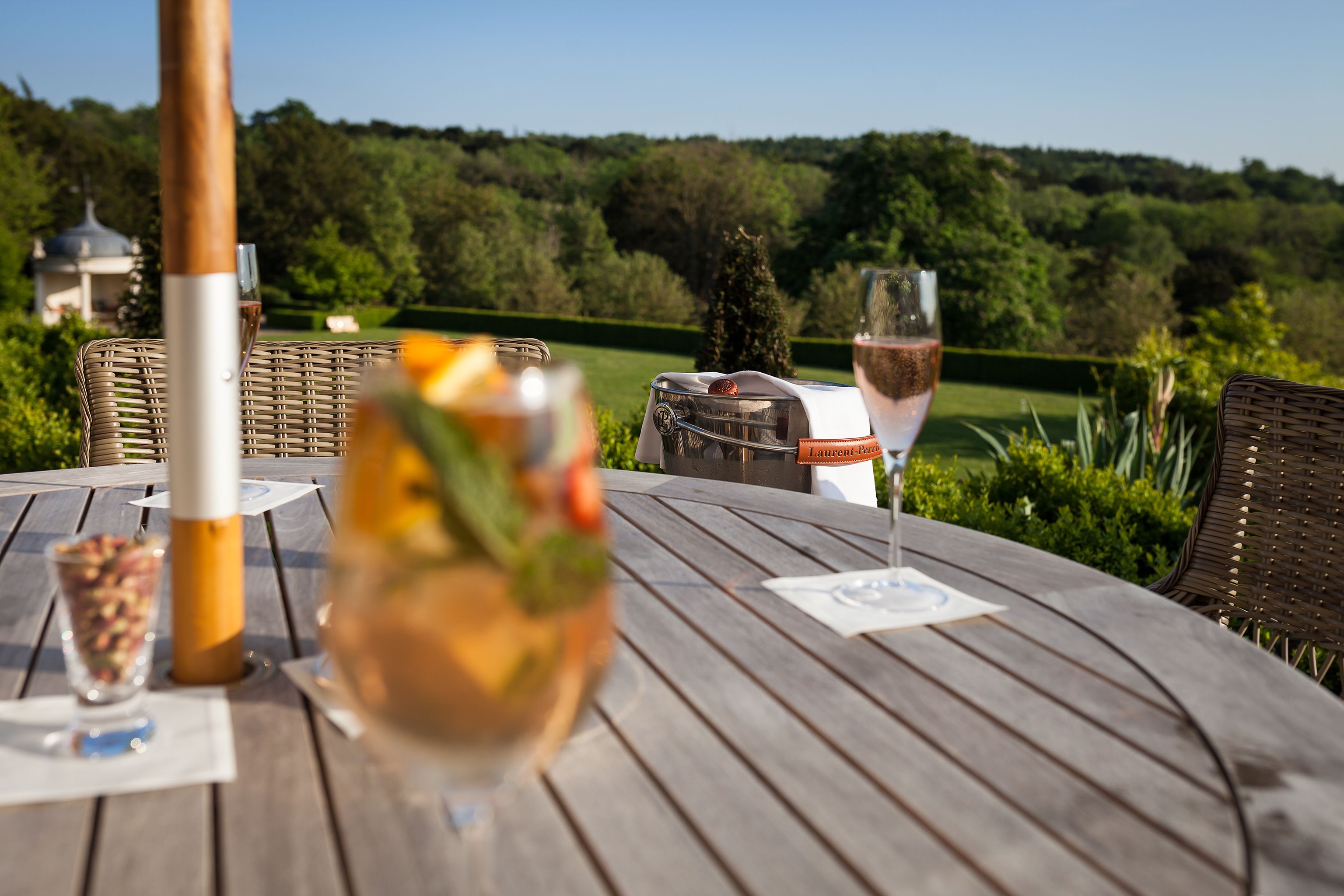 A wooden table with a bottle, a glass of wine, and a glass of water, set against a backdrop of a lush green landscape and a gazebo.