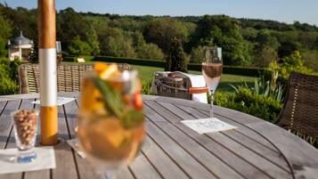 A wooden table with a bottle, a glass of wine, and a glass of water, set against a backdrop of a lush green landscape and a gazebo.