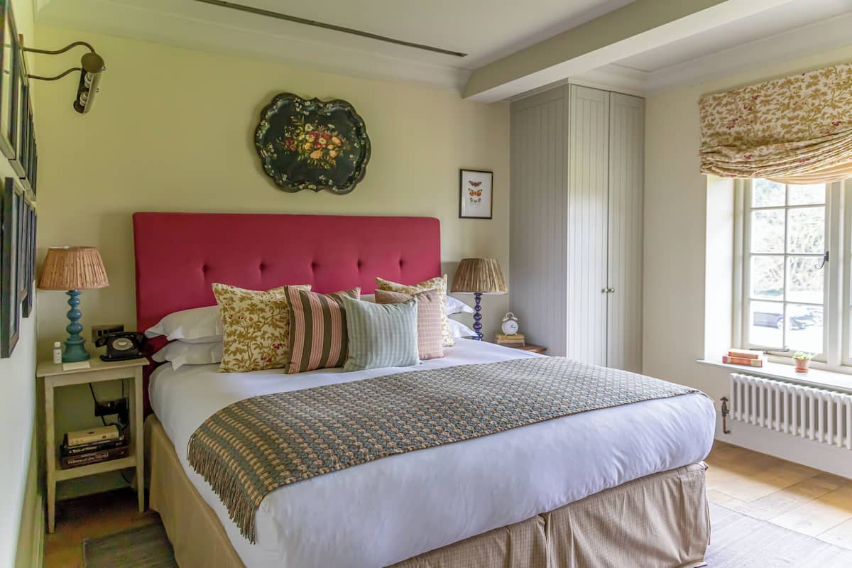A neatly arranged bedroom with a large bed, two bedside tables, a red headboard, a decorative wall piece, and a window with a view.