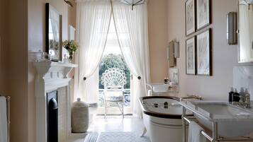 Un baño con bañera, lavabo y espejo. Se ve una ventana con cortinas y vistas al exterior con vegetación.