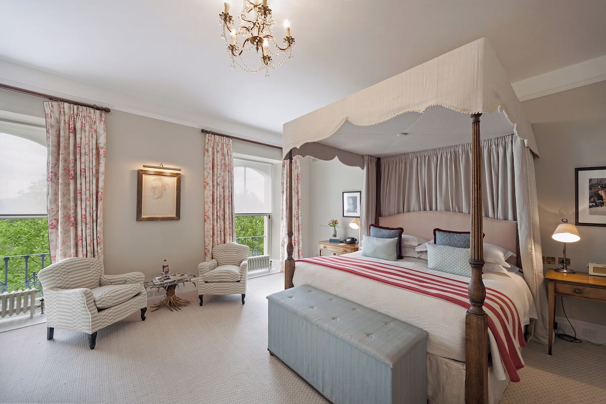 A four-poster canopy bed with a red and white striped bedspread, a wooden bench, two armchairs, a small table, and a nightstand with a lamp.