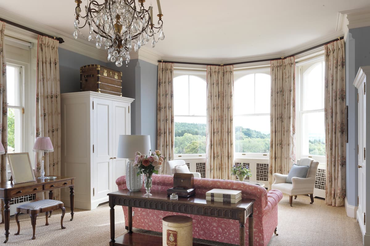 A room with a large window, a chandelier, a pink sofa, a wooden table, and a white cabinet.