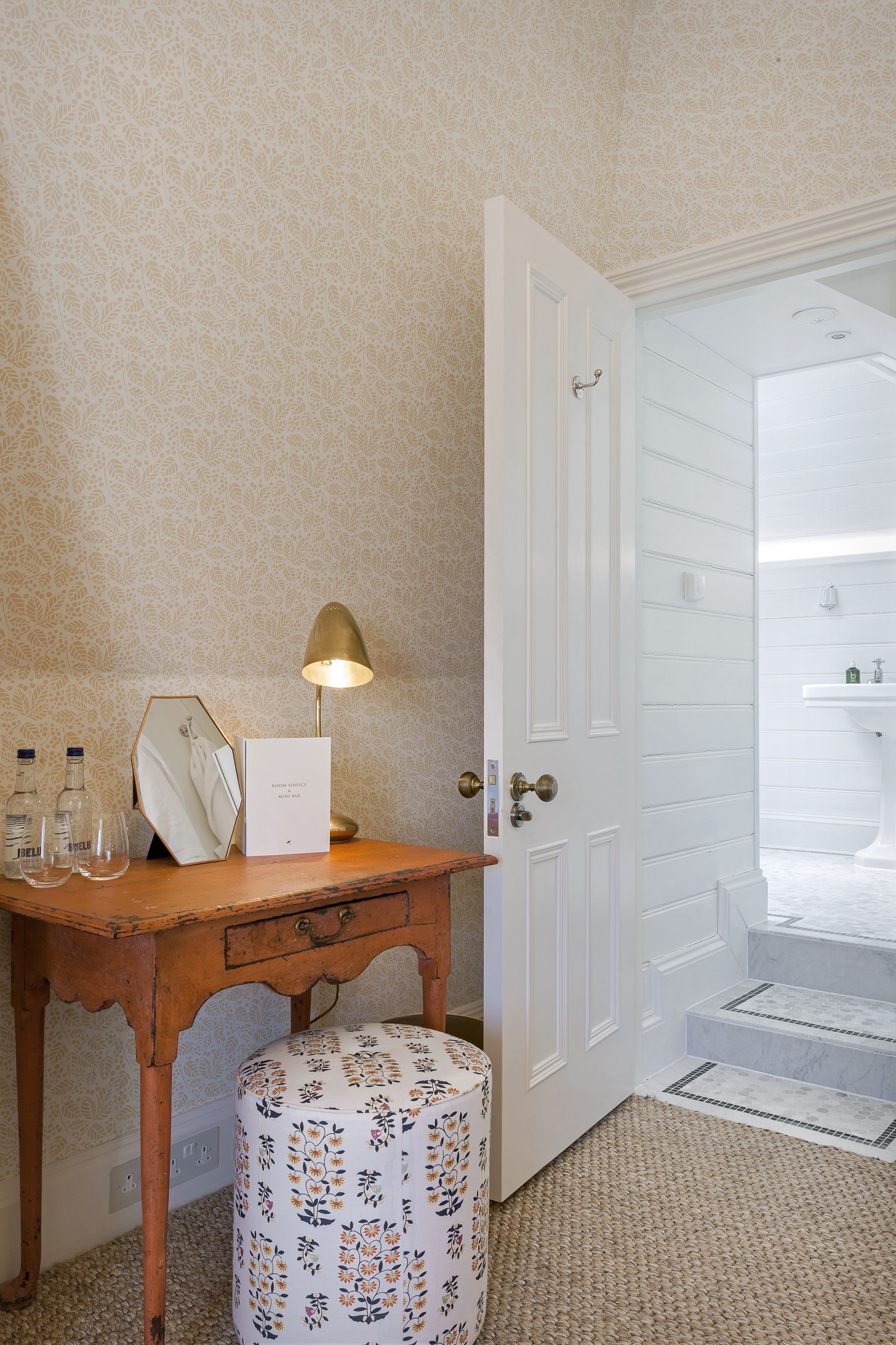 A wooden desk with a lamp, a mirror, and a small stool with a floral pattern. A white door leads to a bathroom with a sink and steps.