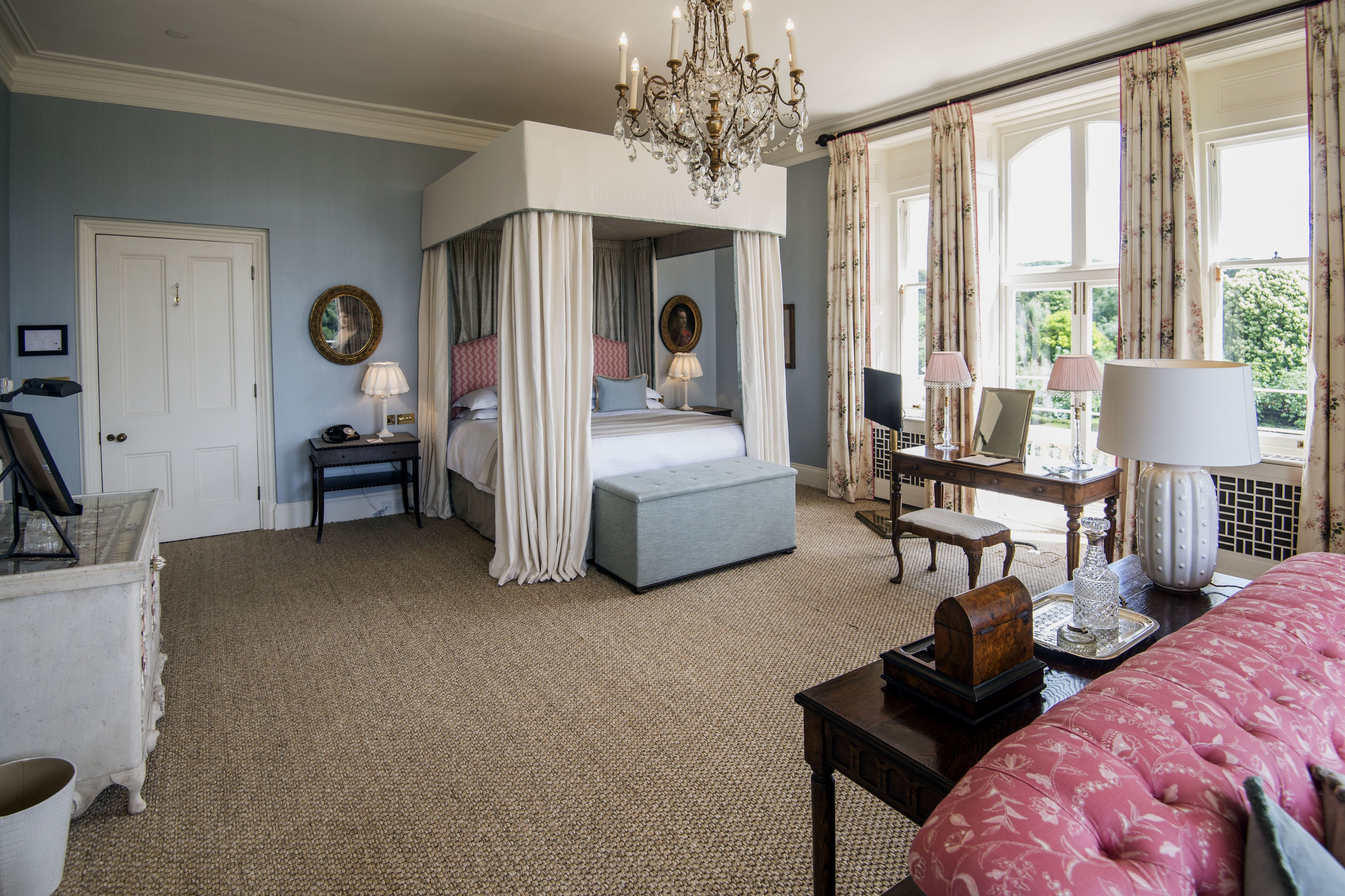 A bedroom with a four-poster bed, a chandelier, and a view of the outdoors through large windows.