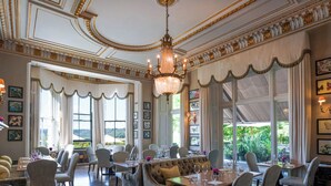 A formal dining room with ornate ceiling details, chandeliers, and large windows offering a view of greenery. - Beaverbrook (Leatherhead)
