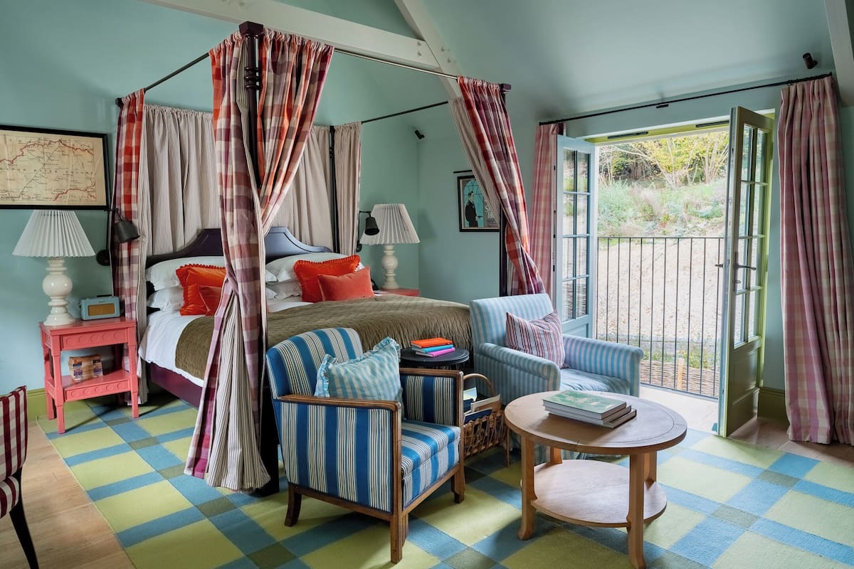 A bedroom with a four-poster bed, a checkered floor, a small table, and a chair.