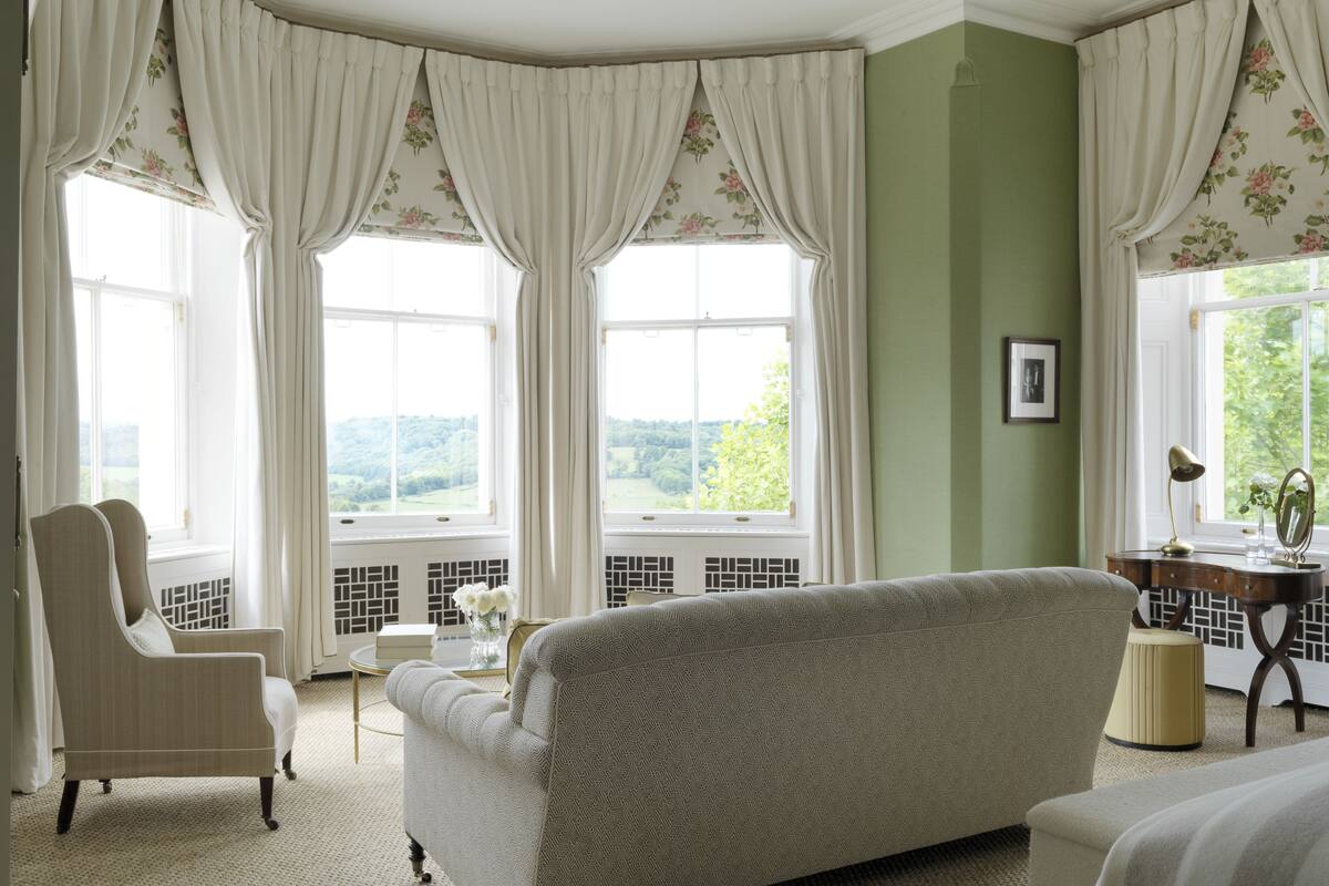 A room with a patterned sofa, a chair, a desk with a lamp, and a view of greenery through large windows with floral curtains.