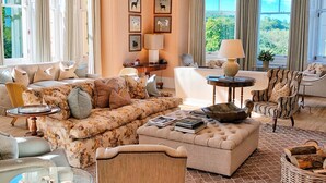 A spacious living room with a large sofa, a coffee table, and a chandelier. There are framed pictures on the wall and a window with curtains. - Beaverbrook (Leatherhead)