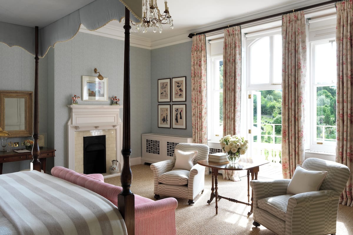 A bedroom with a four-poster bed, a fireplace, a chandelier, and a view of the garden through large windows.