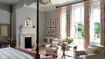 A bedroom with a four-poster bed, a fireplace, a chandelier, and a view of the garden through large windows.