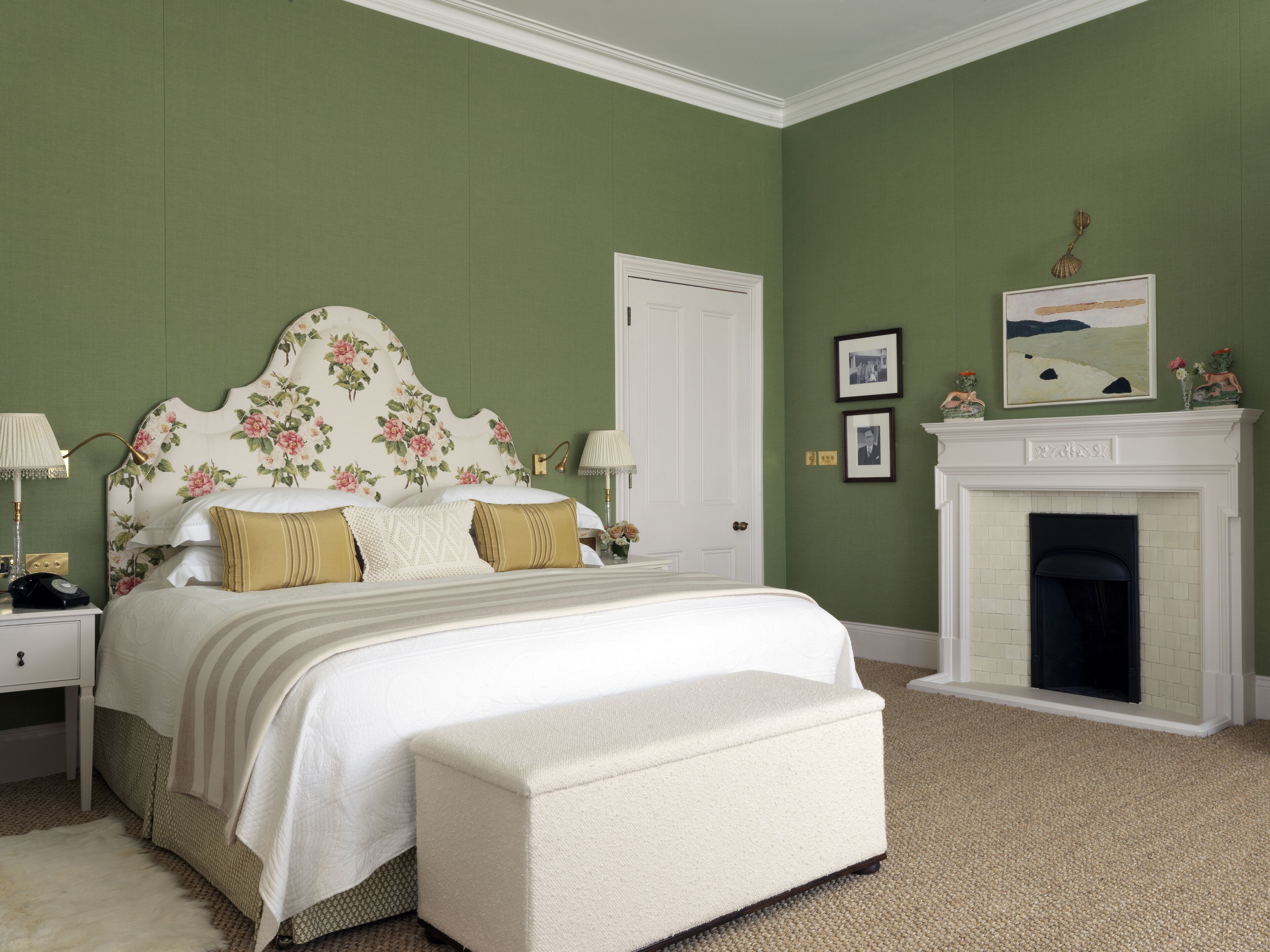 A bedroom with a floral headboard, a white bed, a bench, a fireplace, and green walls.