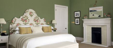 A bedroom with a floral headboard, a white bed, a bench, a fireplace, and green walls.