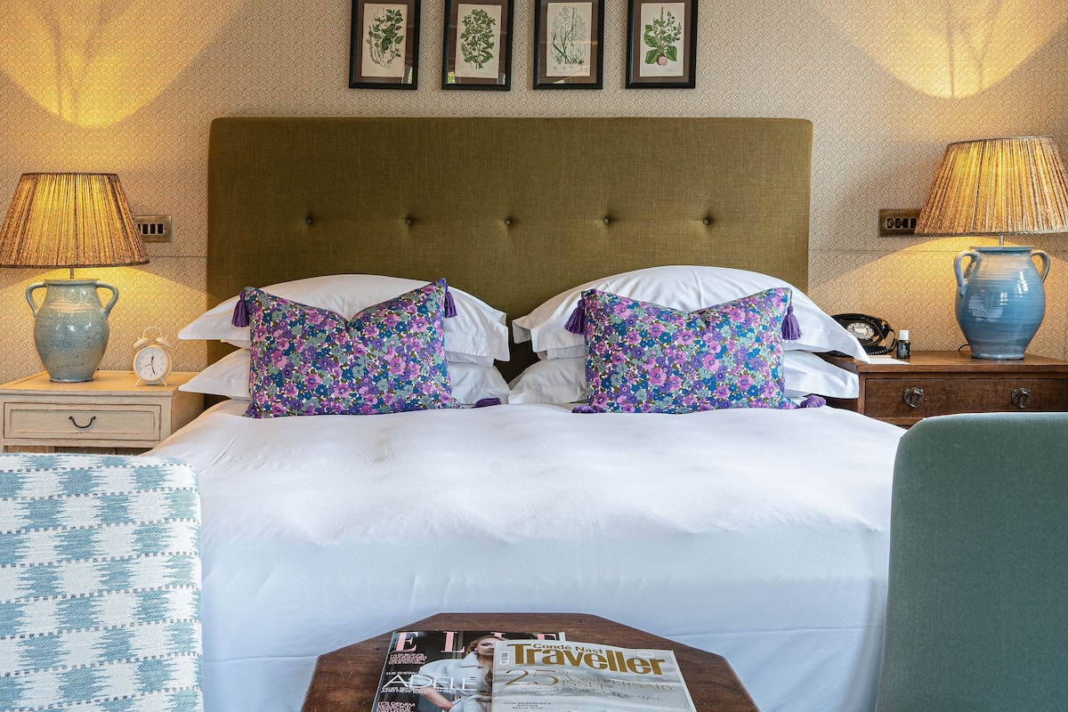 A hotel room with a large bed, two side tables with lamps, a small wooden table with magazines, and a chair.