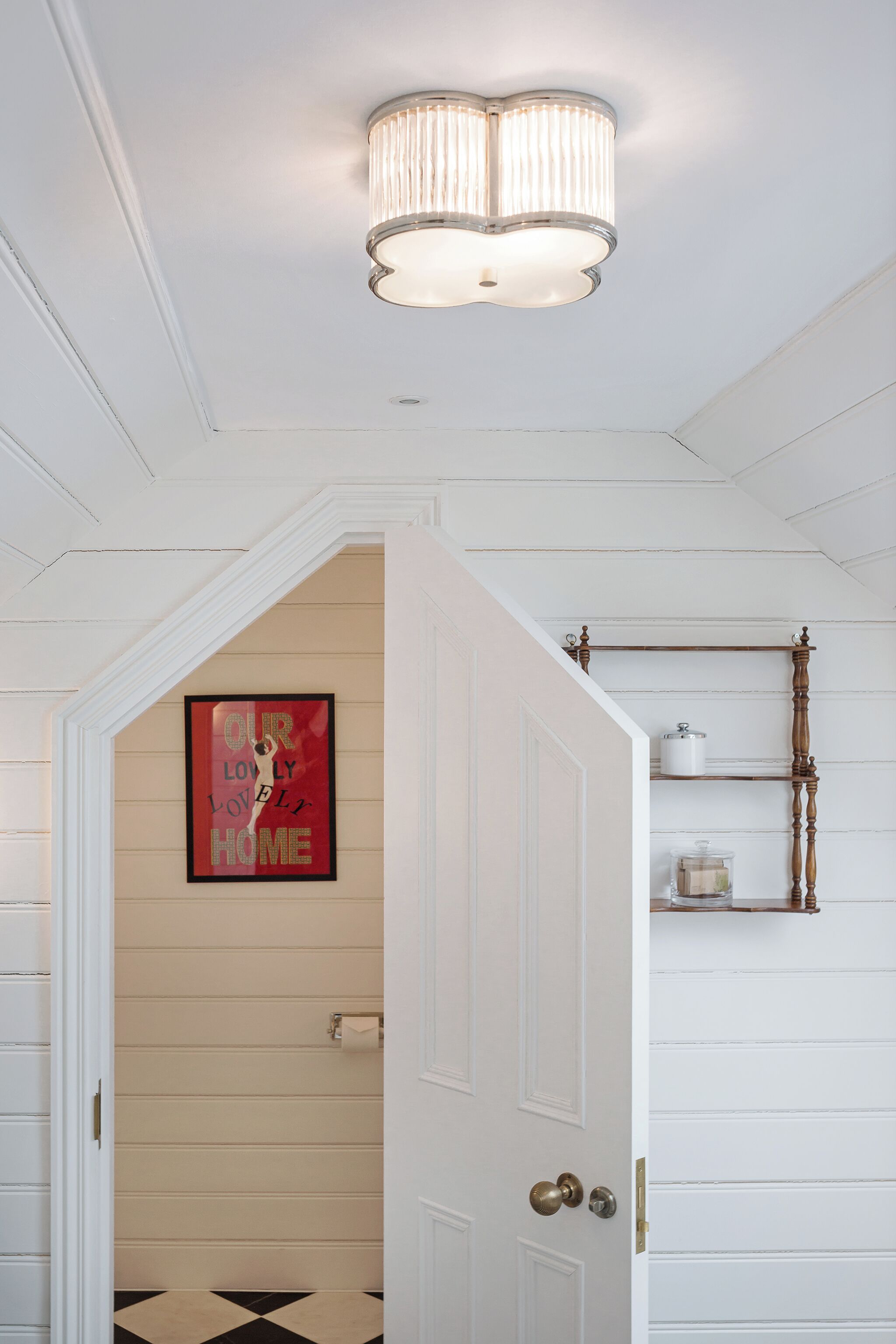 A hallway with a white door, a chandelier, a framed sign on the wall, and a shelf with a container.