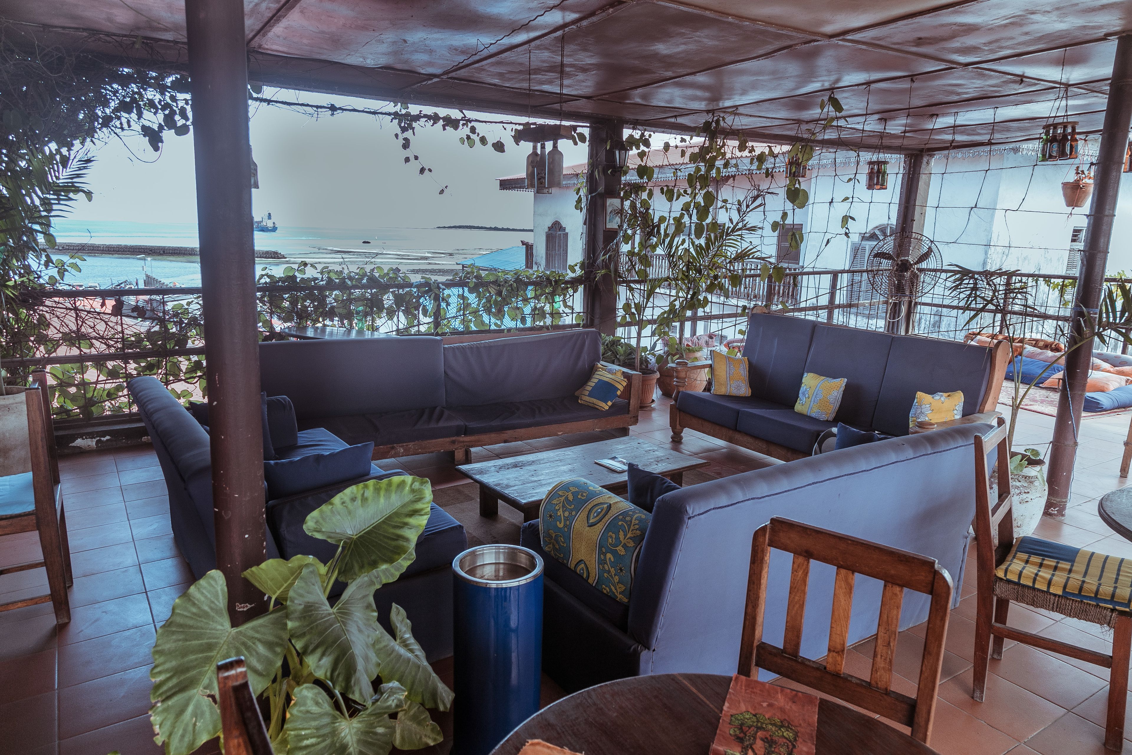 A covered outdoor seating area with a view of the sea, featuring sofas, chairs, and potted plants.