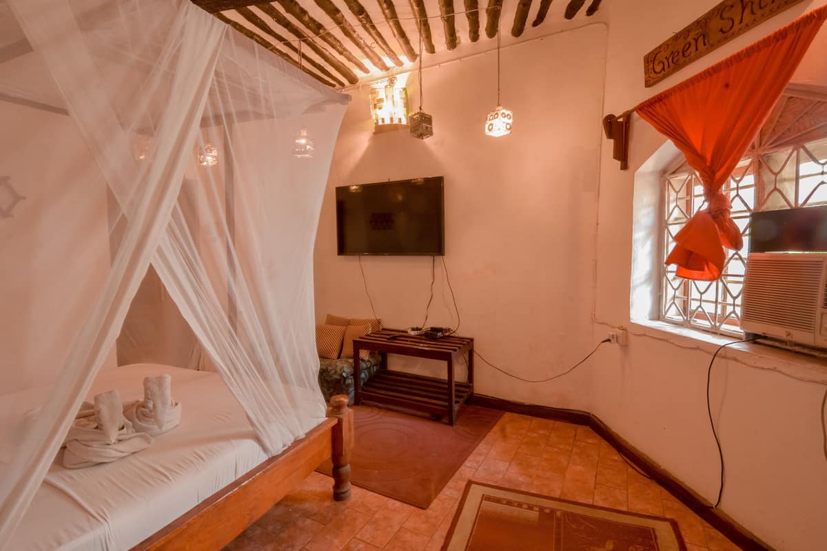 Ein Zimmer mit einem Himmelsbett, einem Fernseher an der Wand, einem kleinen Tisch und einem Fenster mit Klimaanlage.