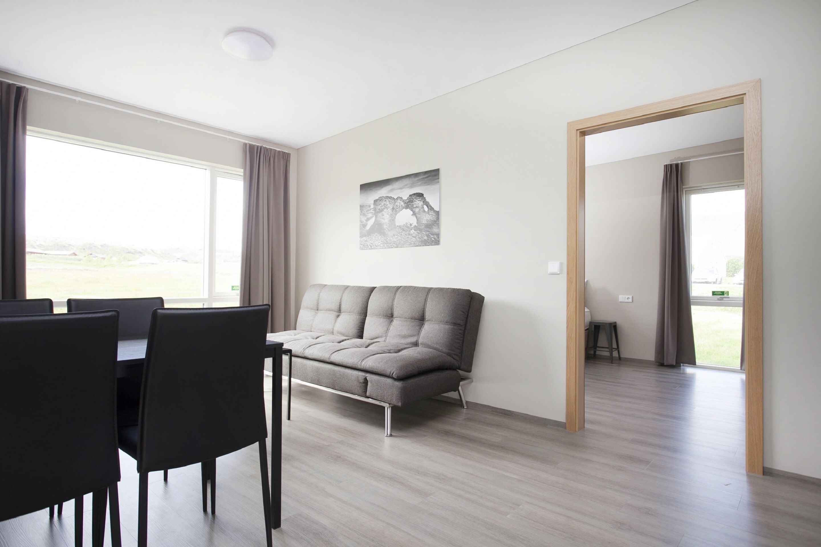 A modern living room with a grey sofa, black chairs, and a dining table. There is a large window with curtains, a painting on the wall, and a wooden door leading to another room.
